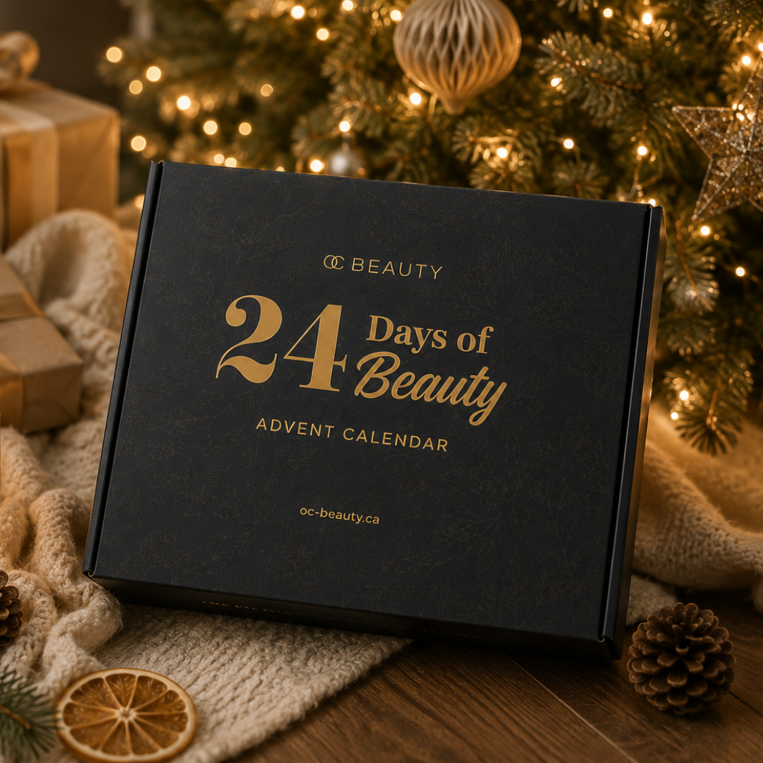 A Navy blue box labelled '24 Days of Beauty Advent Calendar' with gold lettering, placed on a wooden surface with a knitted blanket, pinecones, dried orange slices, and a blurred Christmas tree in the background.