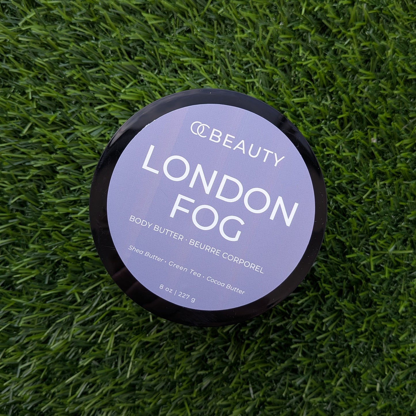 A round black-lidded jar of OC BEAUTY London Fog body butter sits on a textured green artificial grass background.