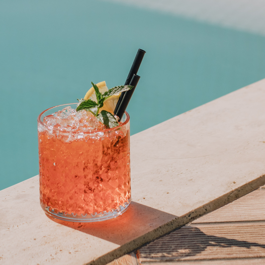 Cocktail with lemon and mint garnish on a wooden surface by a pool.