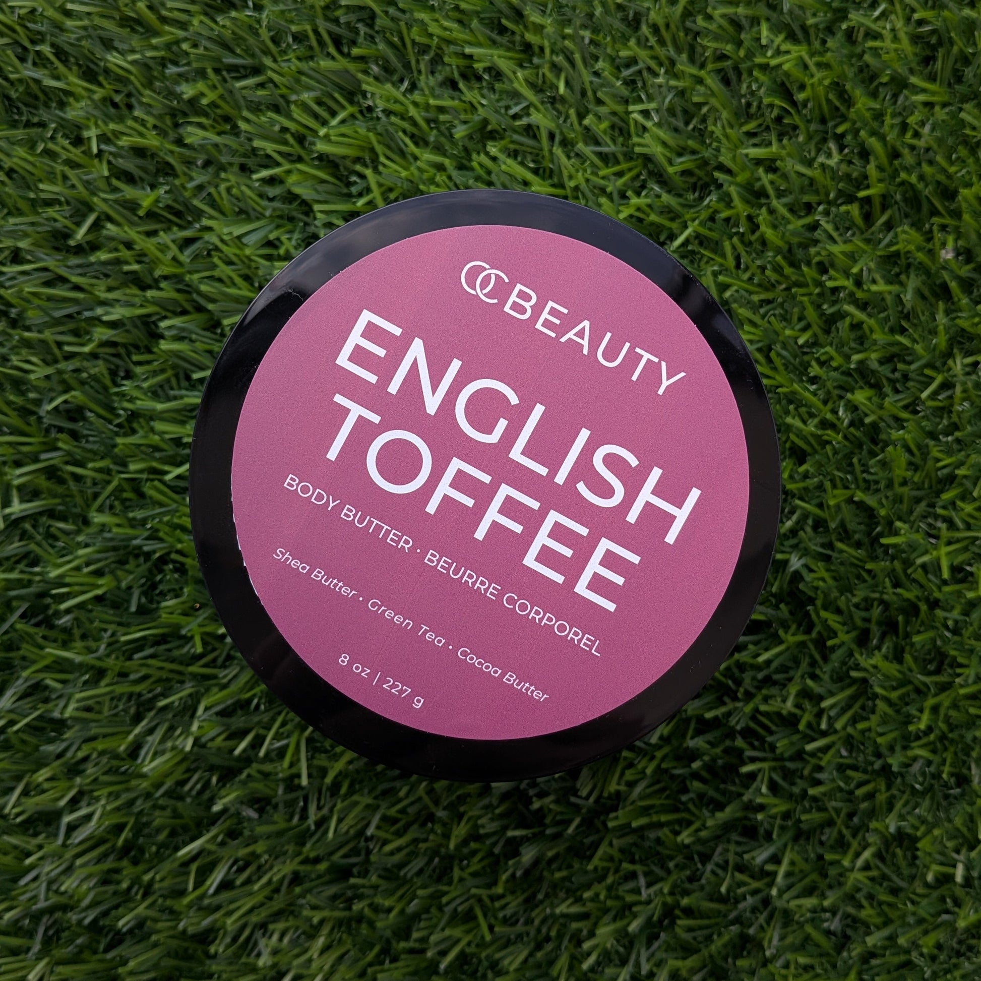 A round black container of OC Beauty English Toffee body butter sits on a textured green grass-like surface. The lid features a pink label with white text.