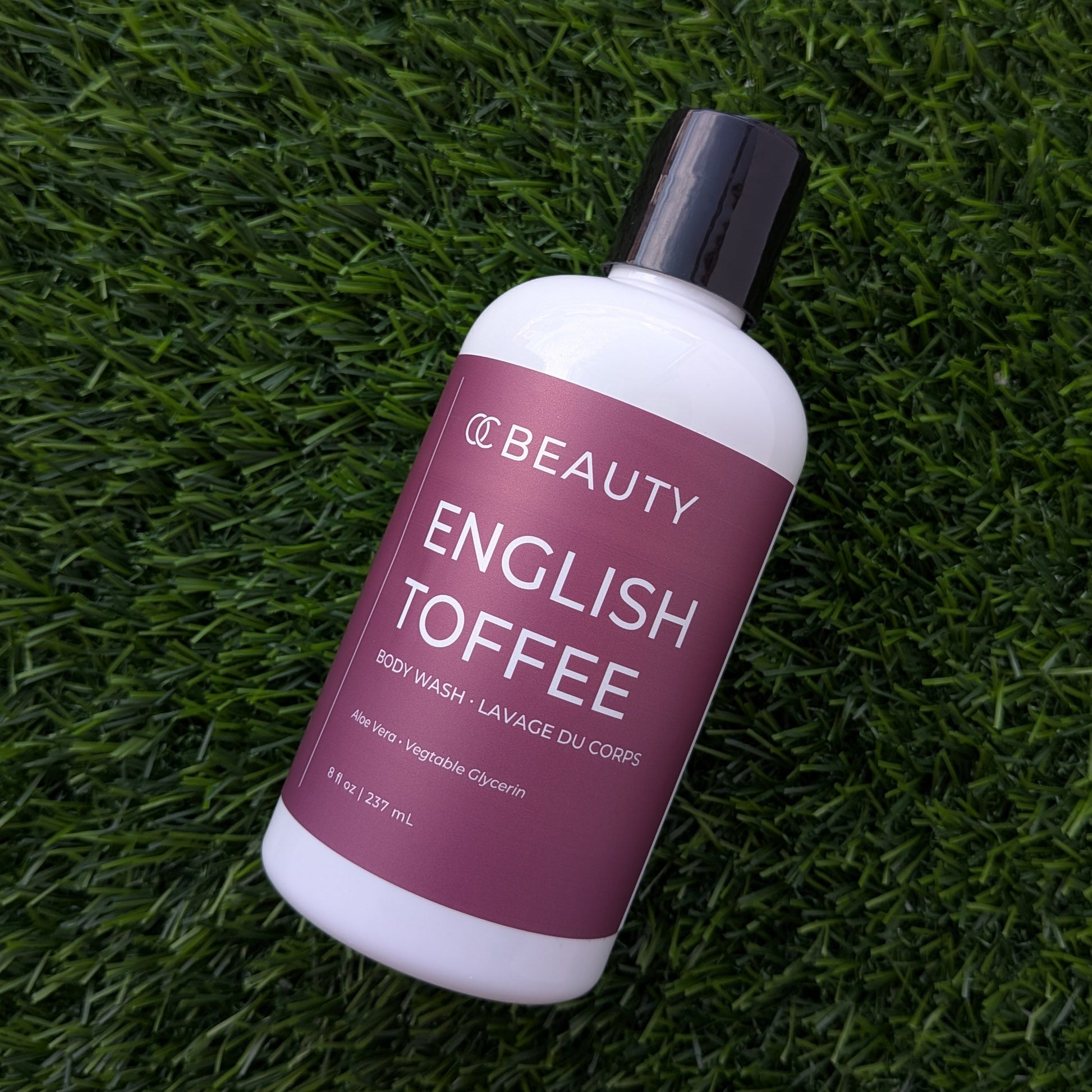 A bottle of OC BEAUTY English Toffee body wash resting diagonally on a textured green grass background.
