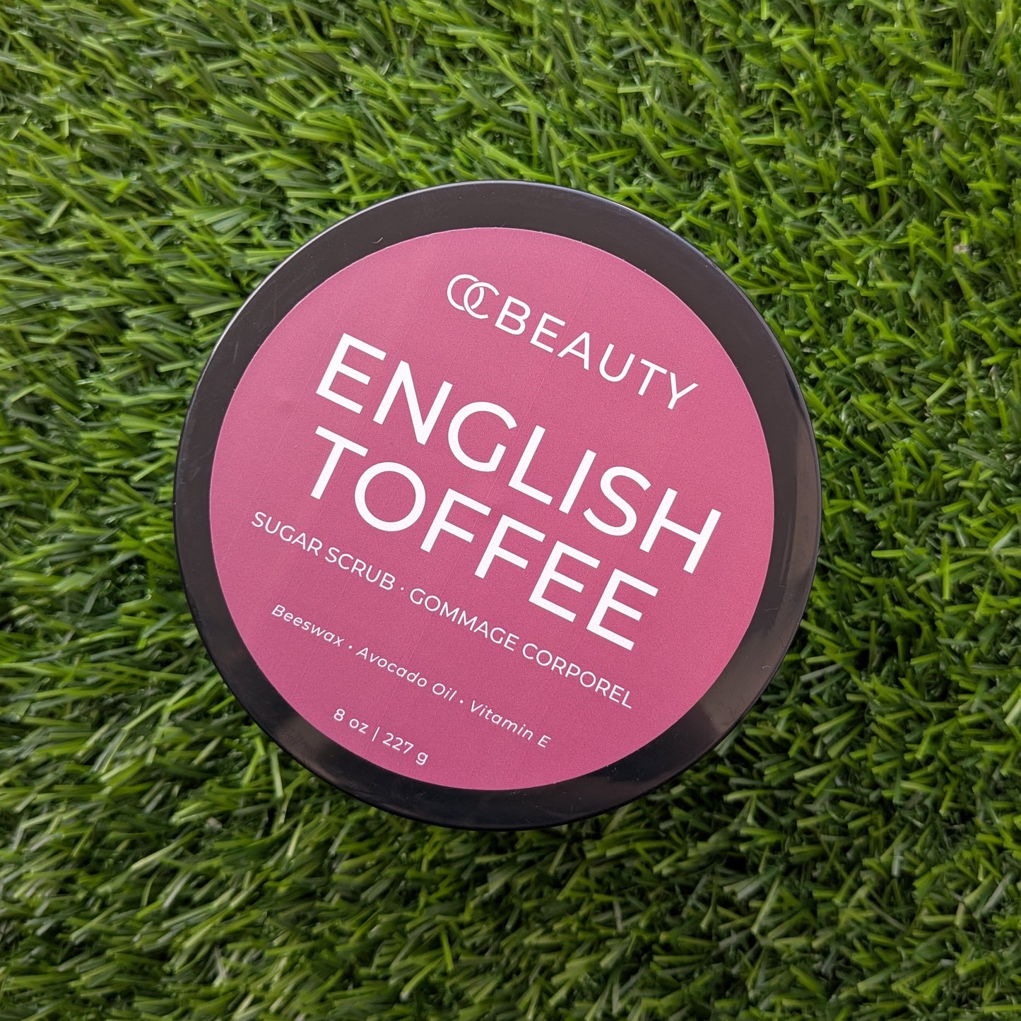 A round black jar lid with a pink label reading 'OC BEAUTY ENGLISH TOFFEE' lies on a green grass-like textured surface.