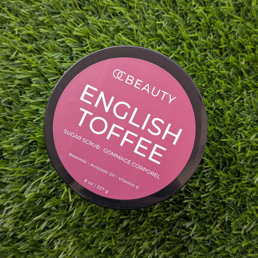 A round black jar lid with a pink label reading 'OC BEAUTY ENGLISH TOFFEE' lies on a green grass-like textured surface.