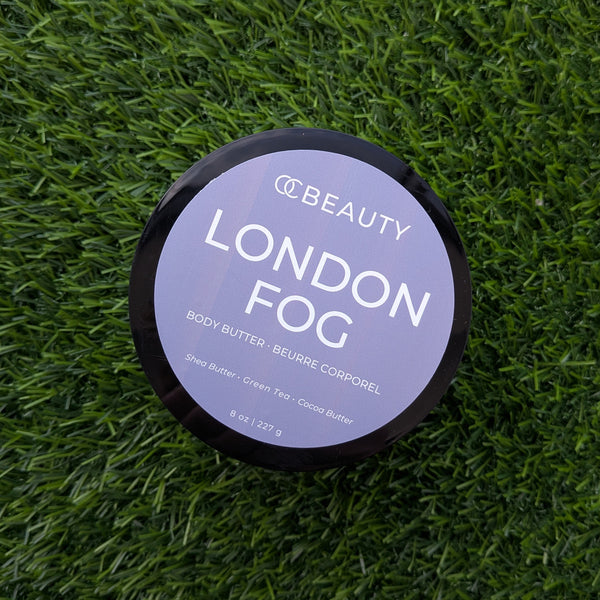 A round black-lidded jar of OC BEAUTY London Fog body butter sits on a textured green artificial grass background.