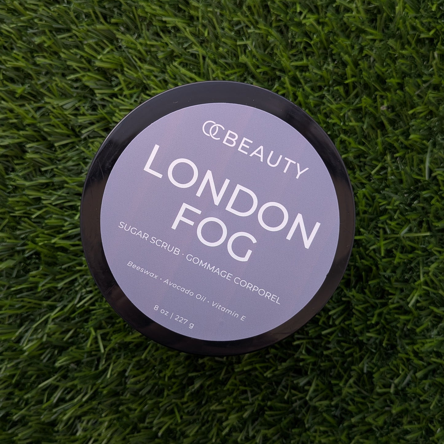 A round black cosmetic tub labeled 'OC BEAUTY LONDON FOG' sits on a textured green grass-like surface. The lid is light gray with white text, and the product appears to be a sugar scrub.