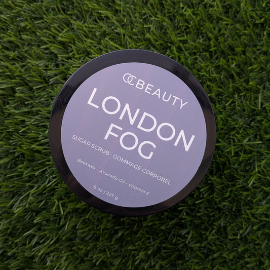 A round black cosmetic tub labeled 'OC BEAUTY LONDON FOG' sits on a textured green grass-like surface. The lid is light gray with white text, and the product appears to be a sugar scrub.