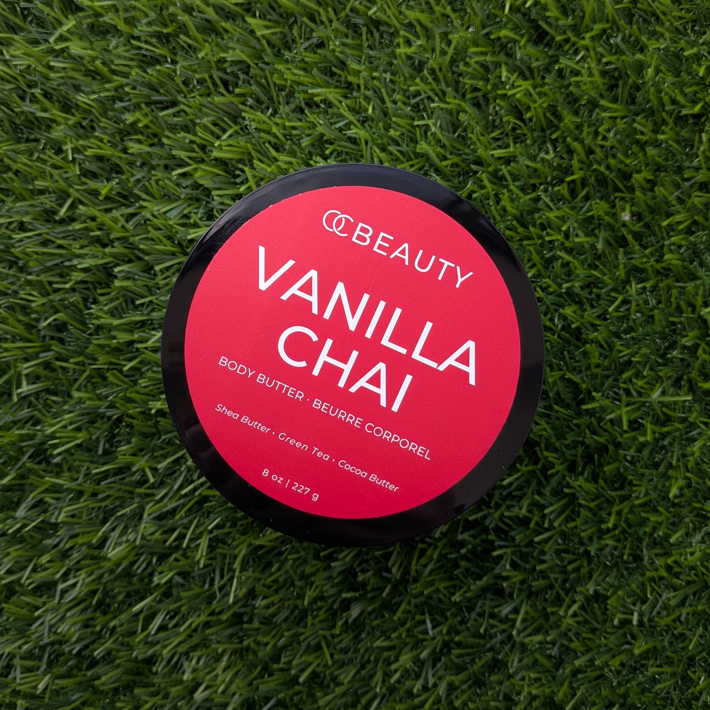 A round black-lidded jar of CC Beauty Vanilla Chai body butter placed on a textured green grass background. The lid features a bright pink-red label with white text.
