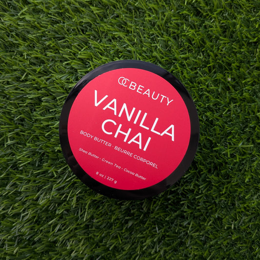 A round black-lidded jar of CC Beauty Vanilla Chai body butter placed on a textured green grass background. The lid features a bright pink-red label with white text.