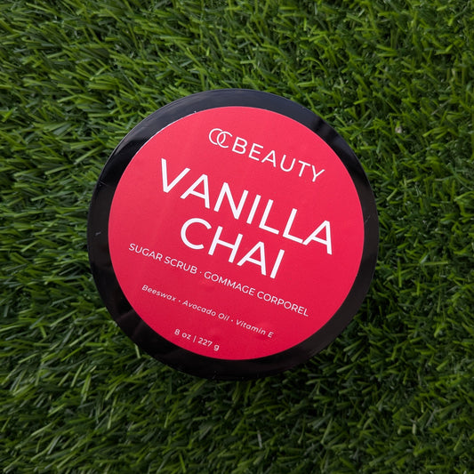 A round black tub of CCBEAUTY Vanilla Chai Sugar Scrub with a bright red label, placed on a green grass background.